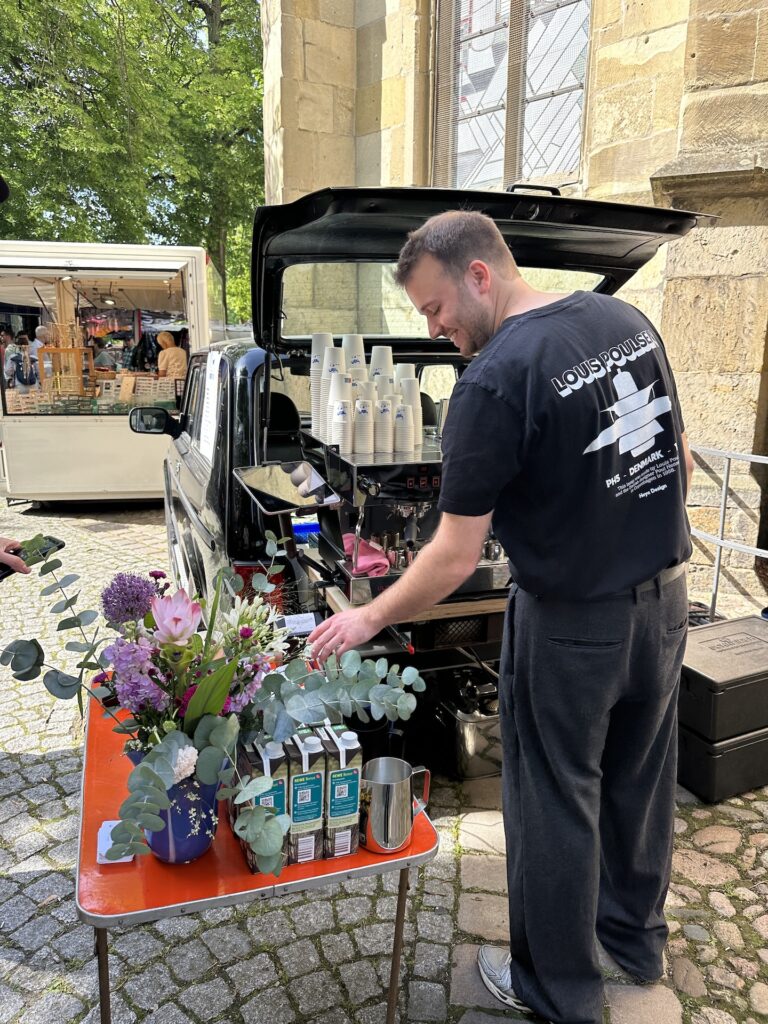 Person vor einem Auto mit einer Kaffeemaschine und greift auf einen Tisch davor