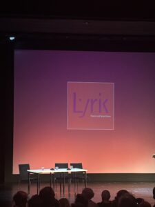 Eine Bühne mit Tisch und Stühlen. Im Hintergrund das Logo des Lyriktreffens an der Wand.